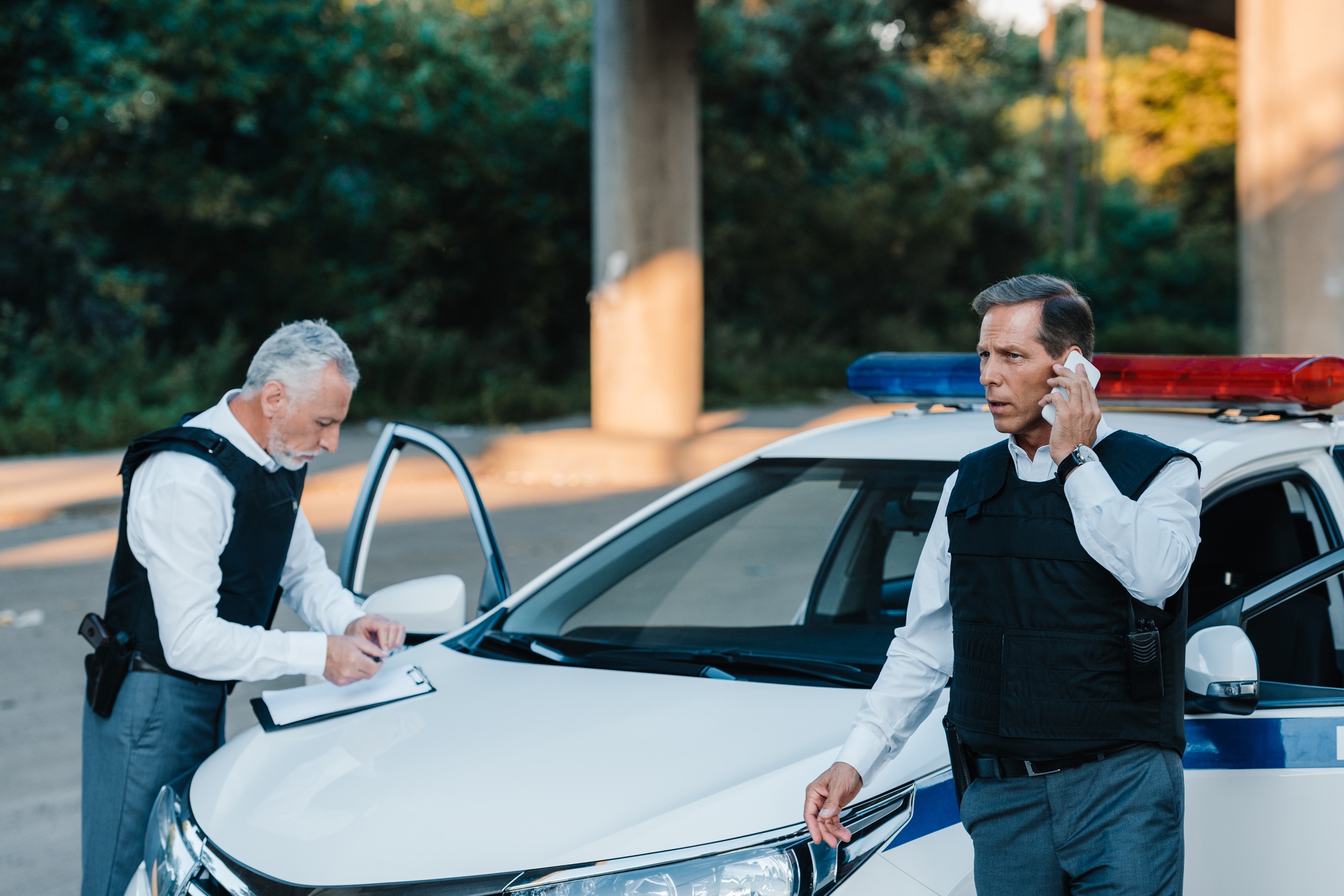 mature police officer talking on smartphone while his colleague writing in clipboard on car at